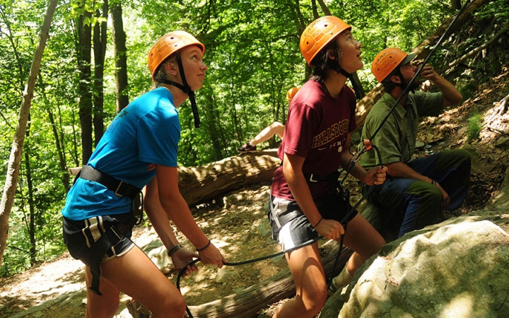 Three people wearing safety gear stand in a wooded area while looking up at an apparent rock climber. Two of them appear to be belaying the climber. 